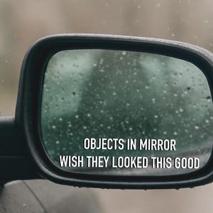 May include: A car side mirror with the text "OBJECTS IN MIRROR WISH THEY LOOKED THIS GOOD". The mirror is black and has water droplets on the glass, with a blurred background.