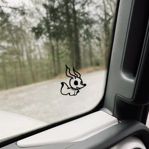 Puede incluir: Una calcomanía de vinilo negro con un diseño de perro de dibujos animados. El perro tiene orejas grandes y un cuerpo pequeño. La calcomanía está adherida a la ventana de un coche, con un bosque borroso y una carretera en el fondo.