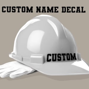 以下が含まれることがあります： 黒い文字で「CUSTOM」と書かれた白い建設用ヘルメット。「CUSTOM NAME DECAL」の文字がヘルメットの上にあります。白い作業用手袋が下にあります。背景は無地のタン色です。