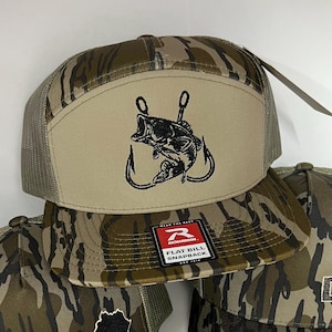 May include: A flat bill snapback hat with a camouflage pattern and a tan front panel. The panel features a black embroidered design of a fish jumping out of the water with two hooks. The hat has a mesh back and a Richardson tag.