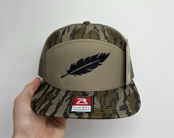 May include: A camouflage flat bill snapback hat with a black feather graphic. The hat features a tan front panel and a mesh back. The brim and top of the hat have a camouflage pattern. The hat has a "Richardson" tag.