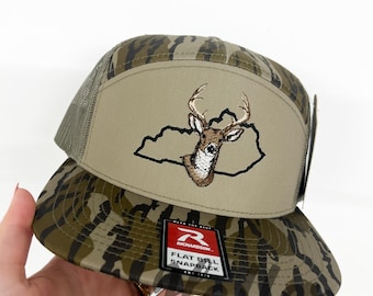 May include: A camouflage flat bill snapback hat with a deer head embroidered on the front. The hat features a tan front panel with a deer head outline and a mesh back. The hat has the text "BOY HATS" on the bottom.