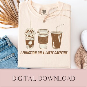 Pode incluir: Camiseta creme com um gráfico de três bebidas de café em tons de castanho. O texto "I FUNCTION ON A LATTE CAFFEINE" está impresso abaixo das xícaras de café. A camiseta tem gola redonda e mangas curtas.