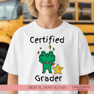 May include: A white short-sleeved t-shirt featuring a graphic of a green frog with a red scarf, a yellow star with the text 4th, and the black text Certified Grader. The design includes small yellow star accents above the frog character.