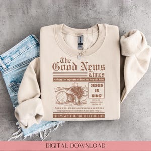 Könnte beinhalten: Beiges Sweatshirt mit dem Text "The Good News Times" in Braun, zusammen mit den Sätzen "Jesus is King!" und "The Way, The Truth, The Life." Das Design beinhaltet eine Illustration eines Grabes und eines Baumes.