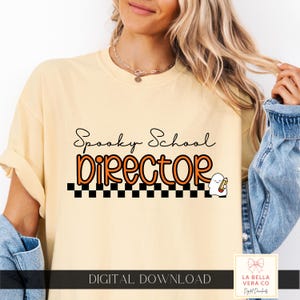 May include: A pale yellow t-shirt featuring the black text Spooky School and the orange text director in a rounded font. Below the text is a black and white checkered pattern and a small white ghost illustration holding a pencil.