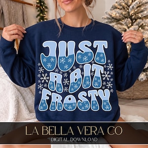 May include: A navy blue crewneck sweatshirt featuring the text "JUST A BIT FROSTY" in a large, bubbly, two-toned light and medium blue font. The letters are decorated with small white snowflake patterns scattered across the entire design.