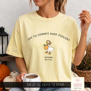 May include: A pale yellow short-sleeve t-shirt featuring the black text OFF TO COMMIT AUNT FOOLERY and AUTUMN EDITION. Between the text is a graphic of a white duck wearing a patterned bonnet and boots, holding an orange pumpkin with green leaves.