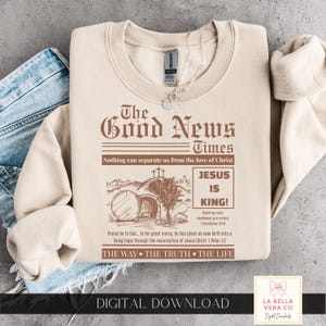 Könnte beinhalten: Ein beigefarbenes Sweatshirt mit Rundhalsausschnitt und einem braunen Grafikdruck, der wie eine Zeitungs-Titelseite mit dem Titel The Good News Times gestaltet ist. Das Design zeigt ein leeres Grab mit drei Kreuzen, den Text Jesus is King und Verse.