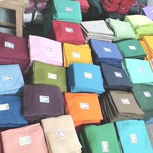 Puede incluir: Exhibición de cuadrados de tela doblados en varios colores, incluyendo rosa, azul, verde y amarillo. Cada cuadrado tiene una pequeña etiqueta blanca. Las telas están apiladas, creando una disposición colorida y organizada.