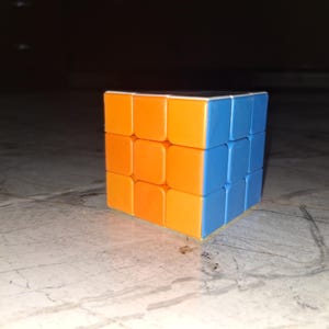 Puede incluir: Un clásico rompecabezas del Cubo de Rubik con lados naranjas y azules. El cubo es un cubo 3x3x3, un juguete rompecabezas popular. Los colores son brillantes y el cubo está enfocado, sobre un fondo oscuro.