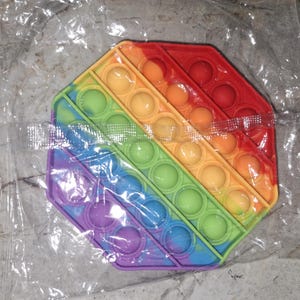 May include: A multi-coloured, octagon-shaped Pop It fidget toy. The silicone toy features rows of bubble-like depressions in red, orange, yellow, green, blue, and purple. The toy is designed to be pushed in and out for sensory play.