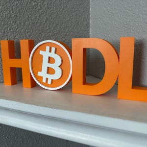 May include: Orange 3D-printed letters spelling "HODL" with a Bitcoin symbol. The Bitcoin symbol is white with an orange border. The letters are displayed on a white shelf against a grey wall.