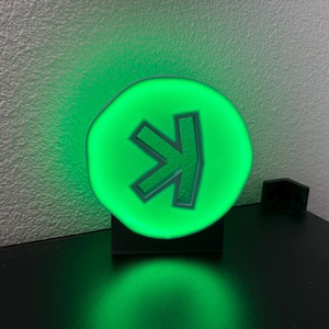 Può includere: Una luce verde circolare con un simbolo a freccia verde scuro. La luce è montata su una base nera, che emette una luce verde. Lo sfondo è una parete bianca strutturata. La luce misura circa 10 cm di diametro.
