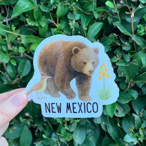 May include: A sticker featuring a brown bear and a bird, with the text "NEW MEXICO" in white. The bear is depicted in a realistic style, and the bird is a light brown color. The sticker is set against a light blue background.