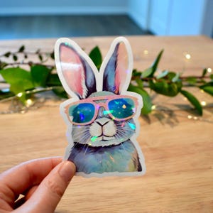 May include: A sticker featuring a cool bunny wearing pink sunglasses. The bunny has blue and gray fur with pink-lined ears. The sticker has a holographic, iridescent finish, and is held by a hand.