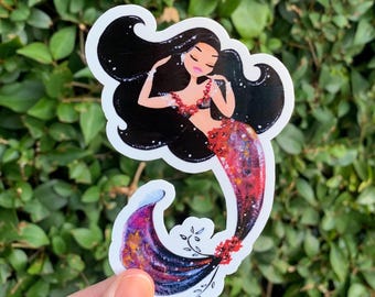 May include: A sticker featuring a mermaid with long black hair, a red and black top, and a red and purple tail. The mermaid is adorned with a red necklace and earrings. The sticker is held by a hand against a green leafy background.