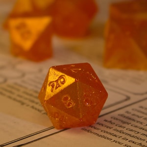 May include: Orange, translucent polyhedral dice set on a printed game sheet. The dice are various shapes, including a 20-sided die in the foreground. A pink marker and purple pen are in the background. The dice are for tabletop role-playing games.