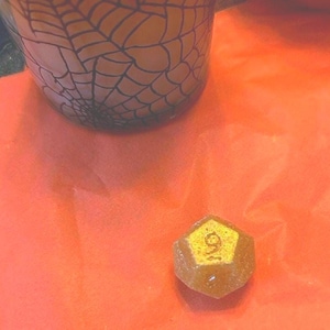 May include: A Halloween-themed image featuring a glass with a black spiderweb design and a small, amber-colored, multi-sided die with the number 9 on it. The die is on an orange surface.