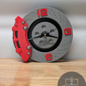 May include: A gray and red brake rotor clock with black clock hands and red numerals at 12, 3, and 6 o'clock. A red brake caliper is attached to the left side. The clock sits on a wooden surface.