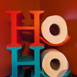 May include: A decorative wall-mounted toilet paper holder with the word "HO HO" in red, orange, and teal. Two rolls of white toilet paper are held in place by teal and orange holders, against a red wall.