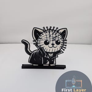 May include: A black and white cat figurine with a Pinhead-inspired design. The cat wears a black coat and has a grid-like pattern on its head, with small pins. The figurine sits on a black base.
