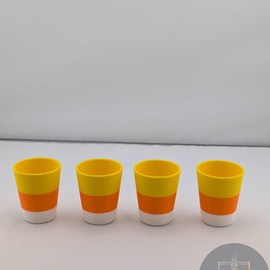 May include: Four shot glasses with a candy corn design. Each glass has a white base, an orange middle section, and a yellow top. The glasses are arranged in a row against a white background.