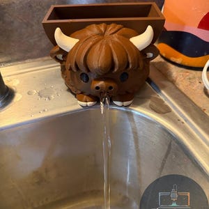 May include: A brown 3D-printed Highland cow-shaped water fountain with white horns and a small rectangular container on its back. Water flows from the cow's nose into a stainless steel sink. The logo "First Layer Fanatics" is visible.
