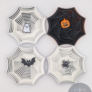 May include: Four Halloween-themed coasters. Two white coasters with a ghost and bat, and two black coasters with a jack-o'-lantern and spider. Each coaster has a layered web design.