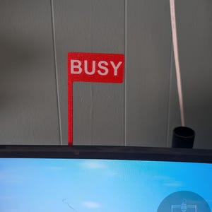 May include: A red sign with the word "BUSY" in white letters. The sign is attached to a thin red post. The sign is placed on top of a computer monitor.