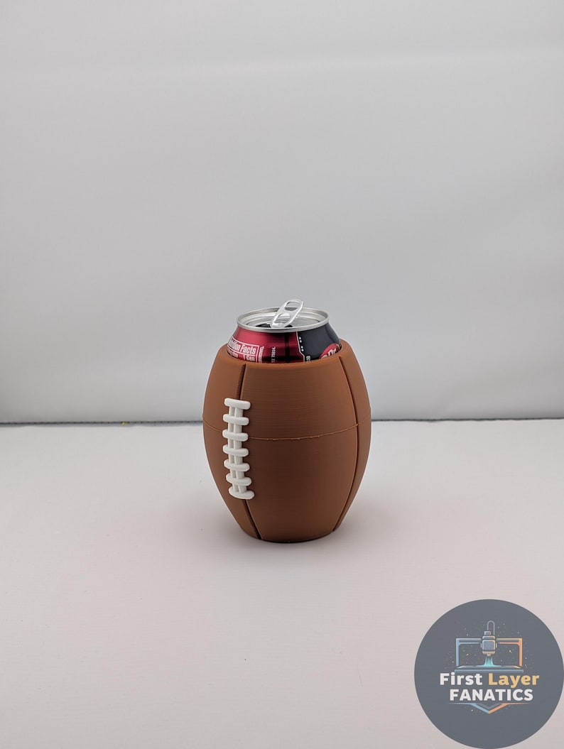 May include: A brown football-shaped can cooler with white stitching detail, holding a red and black can. The cooler is designed to keep beverages cold. The background is plain white.