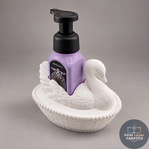 May include: A white swan-shaped soap holder with a woven basket base, containing a square purple soap bottle with a black pump dispenser. The label on the bottle features the text Bath & Body Works and a dark floral graphic design.