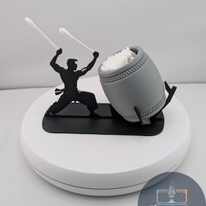 Può includere: Un porta cotton fioc a forma di tamburo grigio con una silhouette nera di un batterista. Il supporto è riempito di cotton fioc bianchi e si trova su una base circolare bianca. L'immagine include il testo "First Layer Fanatics".