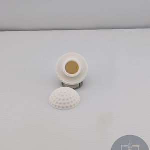 Puede incluir: Un tapón de botella blanco con forma de pelota de golf con una superficie con hoyuelos, junto a una base a juego. El tapón tiene una parte superior cilíndrica con una abertura central. El diseño es simple y moderno.
