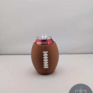 May include: A brown football-shaped can cooler with white stitching detail, holding a red and black can. The cooler is designed to resemble a football. The can has a silver pull tab.