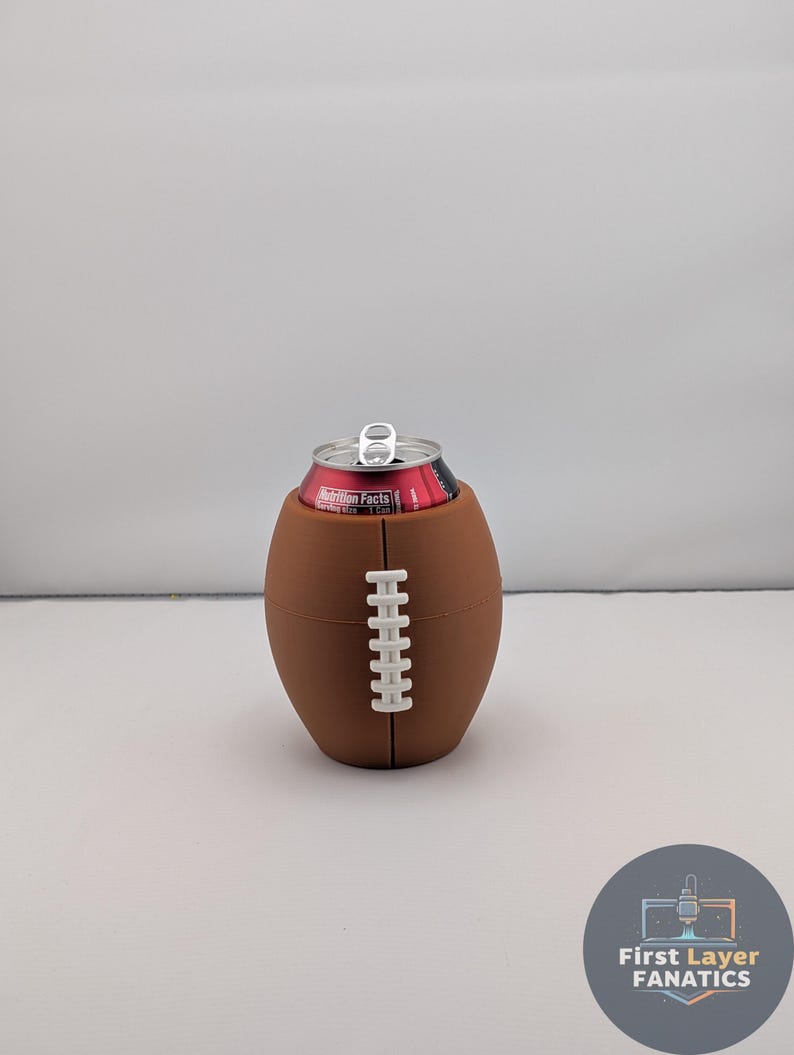 May include: A brown football-shaped can cooler with white stitching detail, holding a red and black can. The cooler is designed to resemble a football. The can has a silver pull tab.