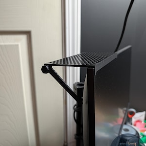 Puede incluir: Un estante de metal negro perforado fijado a un monitor. El estante está soportado por un soporte angular negro. Un cable negro se extiende desde el monitor. El estante parece estar diseñado para guardar objetos pequeños.
