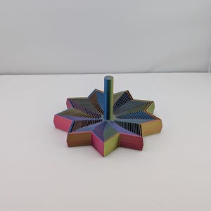 May include: A multi-coloured, star-shaped decorative object with a central post. The object features a layered design with hues of pink, blue, green, and yellow. The star has pointed edges and a cylindrical post extending upwards from the centre.