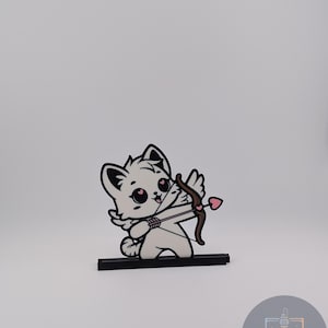 May include: A white cat-shaped figurine with black outlines, holding a bow and arrow with a heart-shaped tip. The figurine is mounted on a black base. The cat has pink cheeks and is in a shooting pose.
