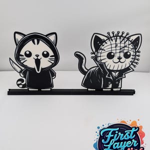 以下が含まれることがあります： 白黒の猫の形をした2つの置物。1匹の猫はナイフを持ったホラー映画のキャラクターに扮しています。もう1匹の猫は別のホラー映画のキャラクターに扮しています。どちらも黒い台座に乗っています。「First Layer Fanatics」のロゴが右下にあります。