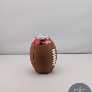 May include: A brown football-shaped can cooler with white stitching detail. A red can with a silver pull tab is inside the cooler. The cooler is on a white surface.