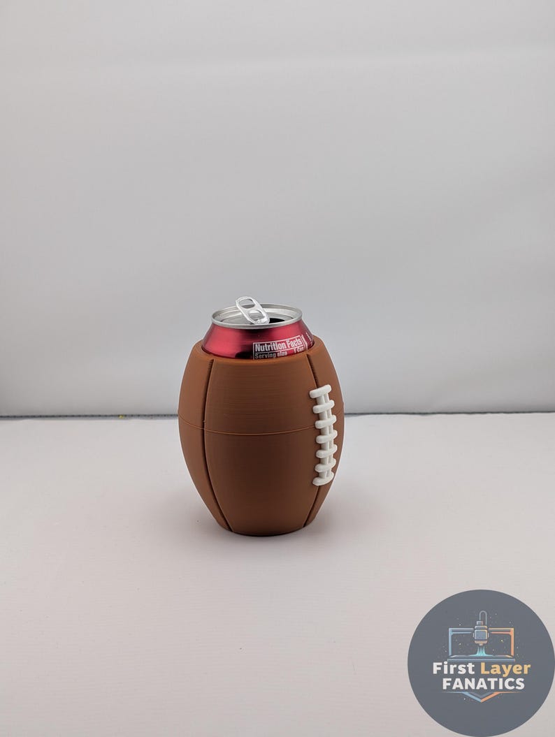 May include: A brown football-shaped can cooler with white stitching detail. A red can with a silver pull tab is inside the cooler. The cooler is on a white surface.