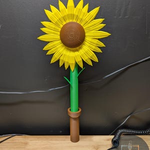 May include: A 3D-printed sunflower with bright yellow petals, a brown center, and a green stem. The sunflower is set in a brown vase-like base. The image is against a dark background.
