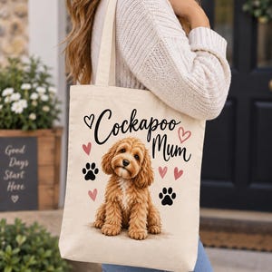 May include: A cream-coloured canvas tote bag featuring a graphic of a brown Cockapoo dog sitting in the centre. The text "Cockapoo Mum" is printed in black script above the dog, surrounded by small pink hearts and black paw print icons.
