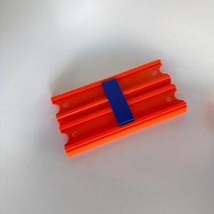 May include: An orange plastic toy track with three parallel grooves and a blue connector piece. The track has rounded cutouts on the sides and small holes. The toy is designed for toy cars.