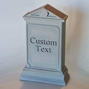 Puede incluir: Un objeto decorativo gris en forma de lápida con las palabras "Custom Text" en la parte delantera. La parte superior de la lápida tiene un pequeño techo con un diseño circular. La base es rectangular.