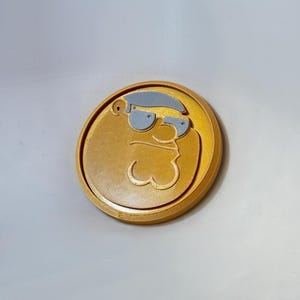 以下が含まれることがあります： 金色の円形メダル。グレーの髪とサングラスをした漫画のキャラクターの顔の3Dプリント画像が特徴です。キャラクターの顔はメダルの中心にあり、薄い灰色の背景があります。