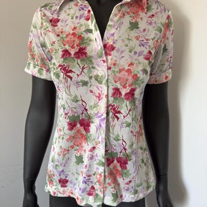 Könnte beinhalten: Kurzärmeliges Hemd mit floralem Muster. Das Hemd hat einen weißen Hintergrund mit einem bunten Aufdruck aus rosa, roten und lila Blumen und grünen Blättern. Das Hemd hat einen Kragen und wird auf einer schwarzen Schaufensterpuppe präsentiert.