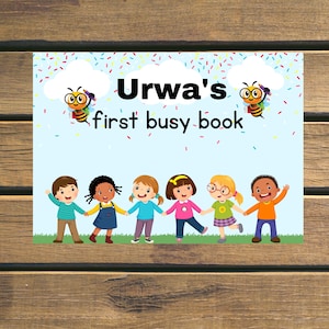 Może przedstawiać: Okładka książki dla dzieci z napisem "Urwa's first busy book". Projekt przedstawia dzieci z kreskówek trzymające się za ręce, pszczoły, konfetti i chmury na jasnoniebieskim tle. Okładka jest umieszczona na drewnianej powierzchni.