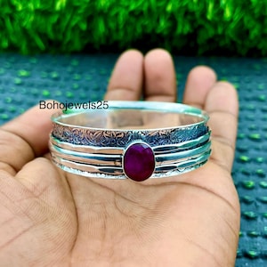 May include: A silver bracelet featuring a large, oval, deep red gemstone. The bracelet has detailed floral patterns and multiple bands. The text "Bohojewels25" is visible on the image.